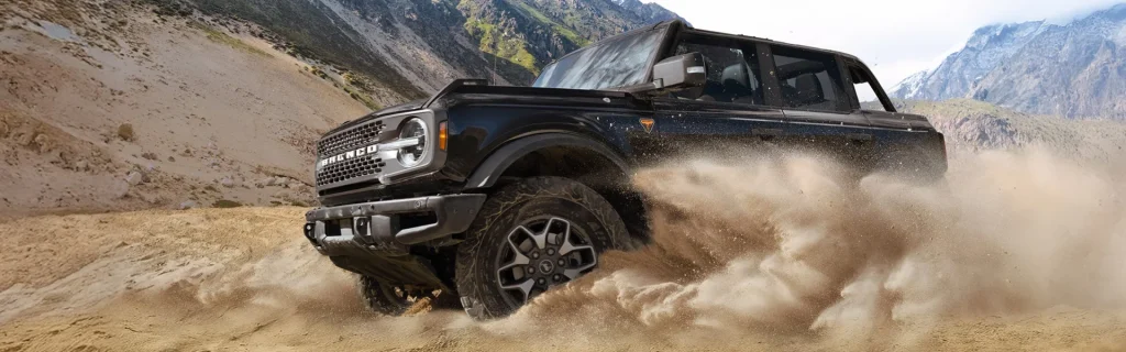Black Ford Bronco off-roading through sand with dust kicking up in a mountainous desert.