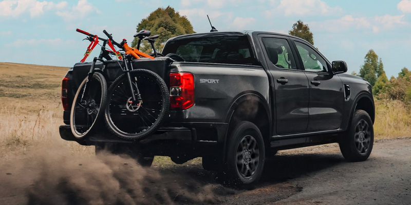 A rear-three-quarter view of a dark grey Ford Ranger Sport kicking up dust on a dirt path, with two mountain bikes secured to a tailgate pad in the truck bed.