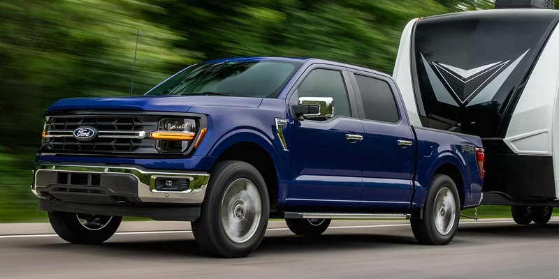 Front-three-quarter view of a dark blue Ford F-150 truck towing a white camper trailer on a highway.