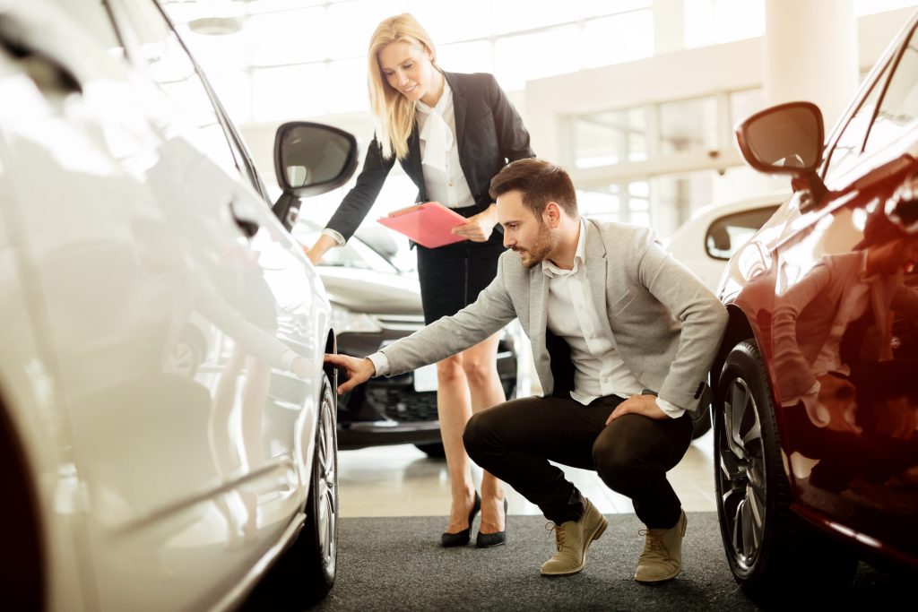 A male customer crouching to inspect a white car wheel while a female sales representative holds a clipboard in a dealership showroom.
